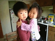 Em uma cozinha, menina oriental sorridente, aparentando 3 anos, abraça forte menino oriental de rosto sério, que aparenta mesma idade.